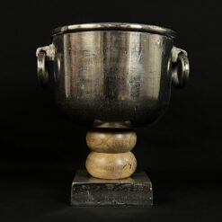Senovinė apvali metalinė vaza su rankenomis tekintu mediniu stovu ant akmens pagrindo.