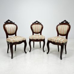Senovinė ąžuolinė kėdė, ornamentais dekoruotu rėmu ir lenktomis kojomis, aptraukta šviesiu dryžuotu audiniu.
